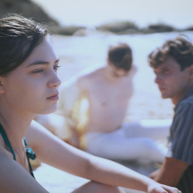 Eine junge Frau sitzt am Strand, im Hintergrund zwei Männer.