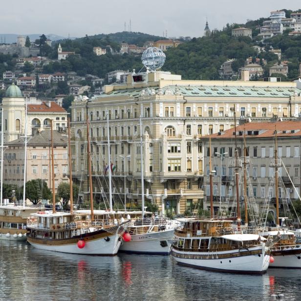 Mehrere historische Segelboote liegen im Hafen von Triest vor der Stadtkulisse.