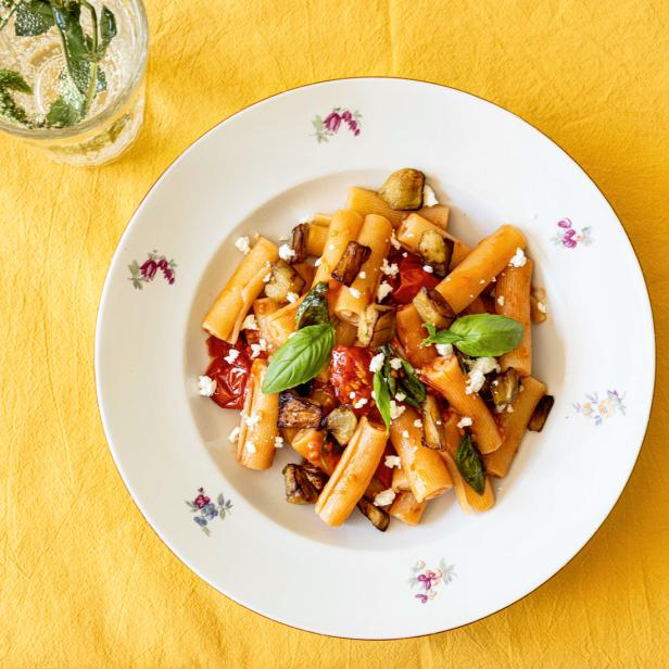 Ein Teller Pasta mit Auberginen, Tomaten, Basilikum und Feta auf einem gelben Tisch.
