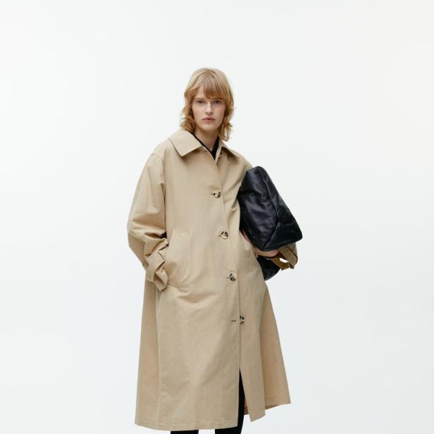 Eine Frau mit blonden Haaren trägt einen beigen Trenchcoat, Leggings und eine schwarze Tasche.