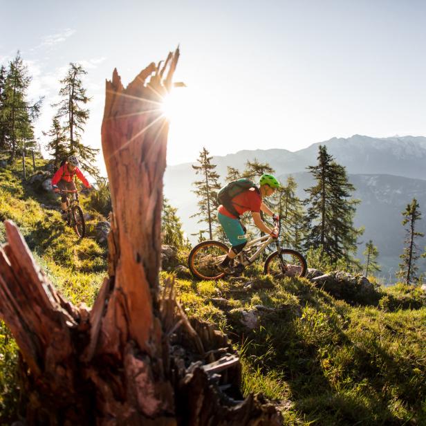 Zwei Mountainbiker fahren auf einem Bergpfad mit Blick auf die Alpen.