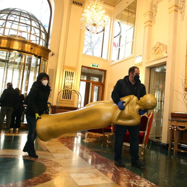 Zwei Personen tragen eine übergroße goldene Oscar-Statue in einem eleganten Gebäude.