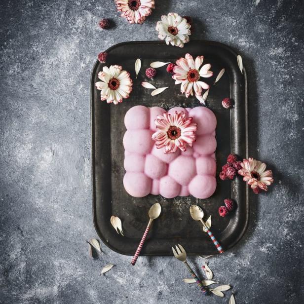 Ein rosa Dessert mit Blüten und Himbeeren auf einem dunklen Serviertablett.