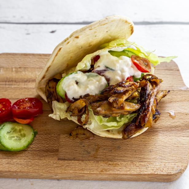 Ein Pita-Sandwich mit Fleisch, Salat, Tomaten, Gurken und Soße auf einem Holzbrett.