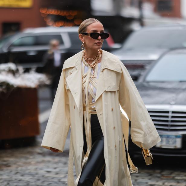 Eine Frau mit Sonnenbrille und Trenchcoat geht auf einer Straße entlang.