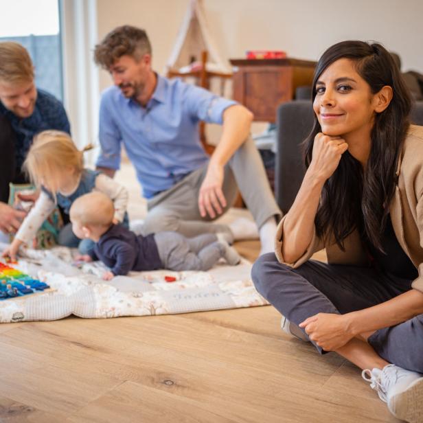 Eine Frau sitzt auf dem Boden, während zwei Männer mit Kindern auf einer Decke spielen.