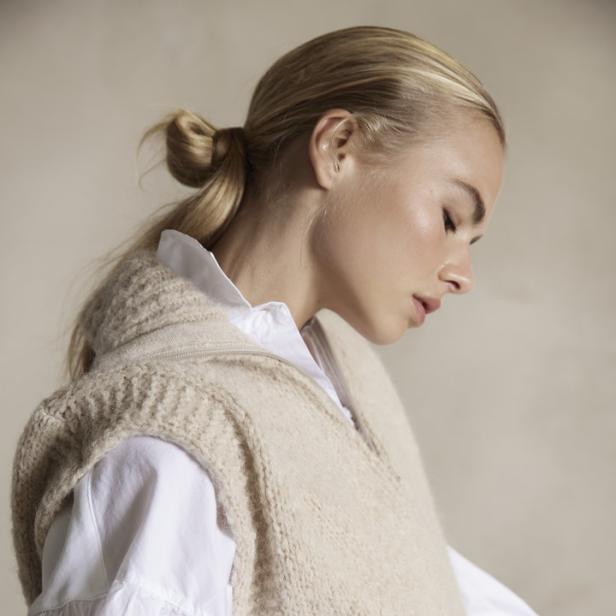 Eine blonde Frau trägt eine weiße Bluse unter einer beigen Strickweste.