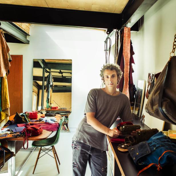 Ein Mann mit lockigem Haar steht in einem Atelier mit Lederwaren und Werkzeugen.