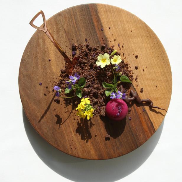 Ein Dessert mit essbarer Erde, Blüten und einer roten Kugel auf einem Holzbrett angerichtet.