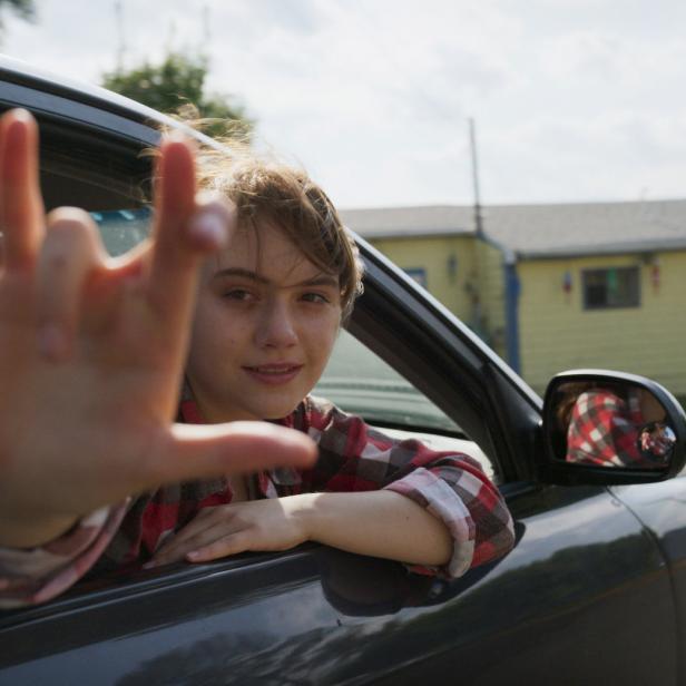 Eine junge Frau lehnt aus einem Auto und formt mit der Hand eine Geste.
