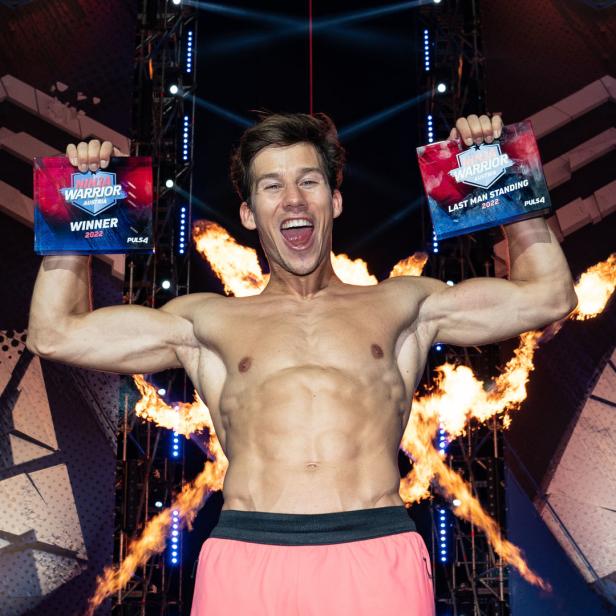 Ein muskulöser Mann jubelt mit zwei „Ninja Warrior Austria“-Trophäen in der Hand vor einer Feuerwand.
