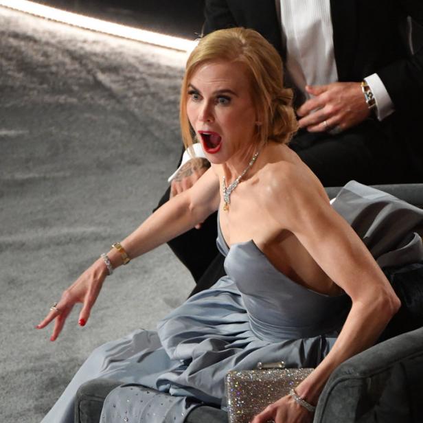 Nicole Kidman sitzt mit offenem Mund und erhobenen Händen auf einem Sofa.