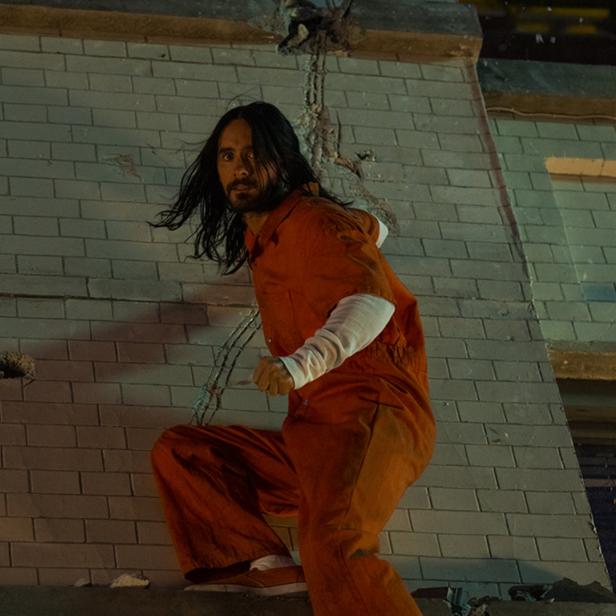 Jared Leto in oranger Gefängniskleidung klettert an einer beschädigten Ziegelmauer hoch.