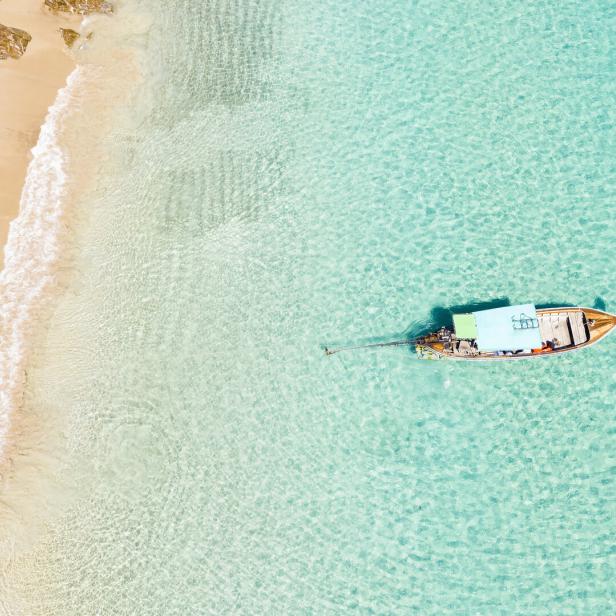 Luftaufnahme von zwei Personen an einem tropischen Strand mit weißem Sand, türkisfarbenem Wasser und einem traditionellen Longtail-Boot. Banana Beach, Phuket, Thailand.