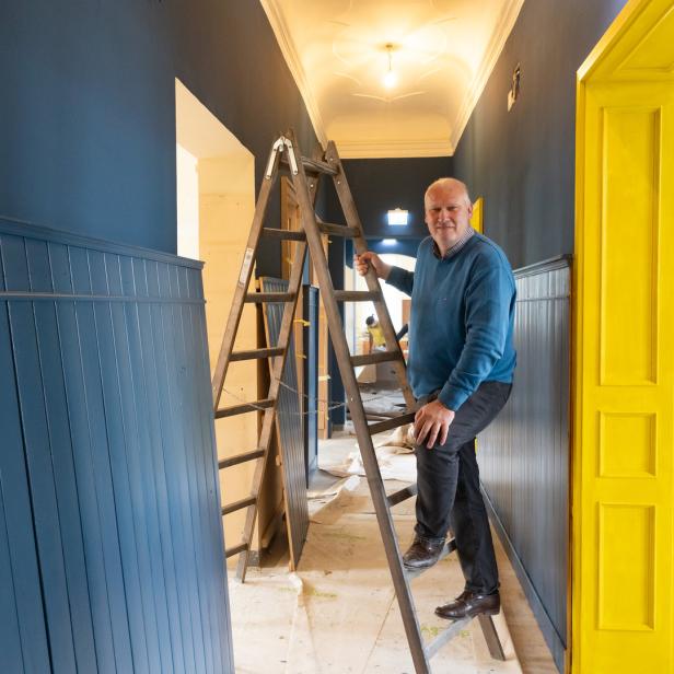 Ein Mann steht auf einer Leiter in einem Flur mit blau gestrichenen Wänden und einer gelben Tür.