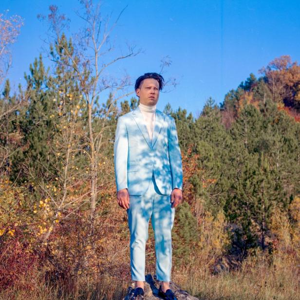 Ein Mann im hellblauen Anzug steht auf einem Felsen in einer herbstlichen Landschaft.