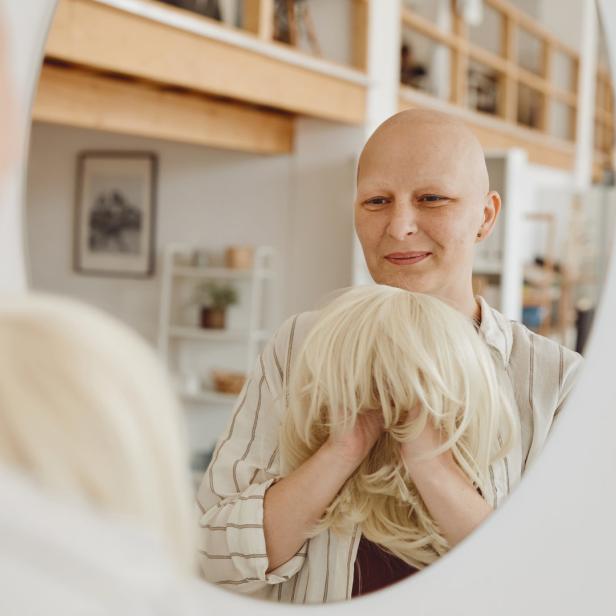 Eine Frau ohne Haare hält eine blonde Perücke vor einem Spiegel.