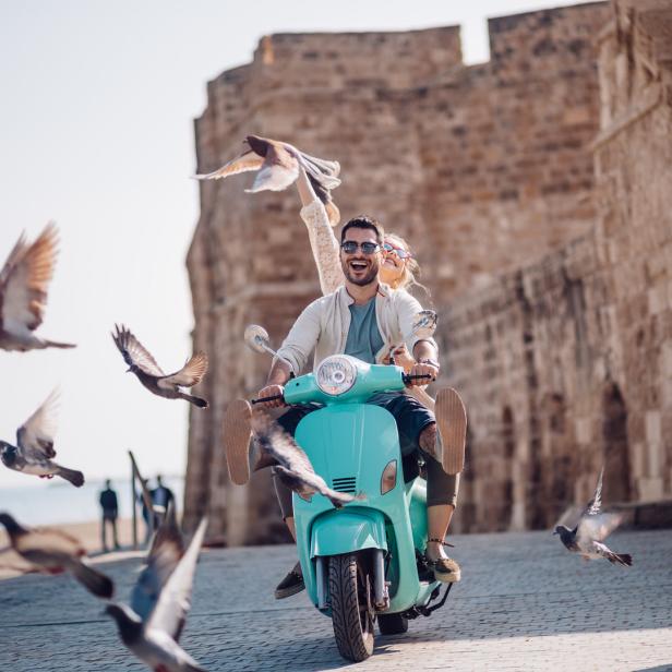 Ein Paar fährt auf einem türkisfarbenen Motorroller, umgeben von fliegenden Tauben.