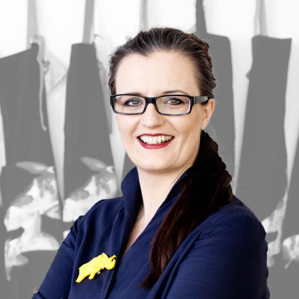 Eine lächelnde Frau mit Brille und einem gelben Elefanten-Anstecker vor einem grauen Hintergrund.
