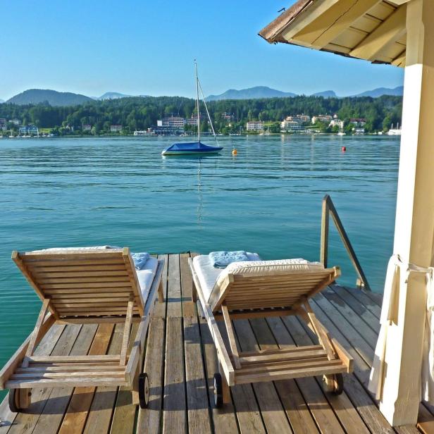 Zwei Liegestühle stehen auf einem Holzsteg mit Blick auf einen See und bewaldete Hügel.