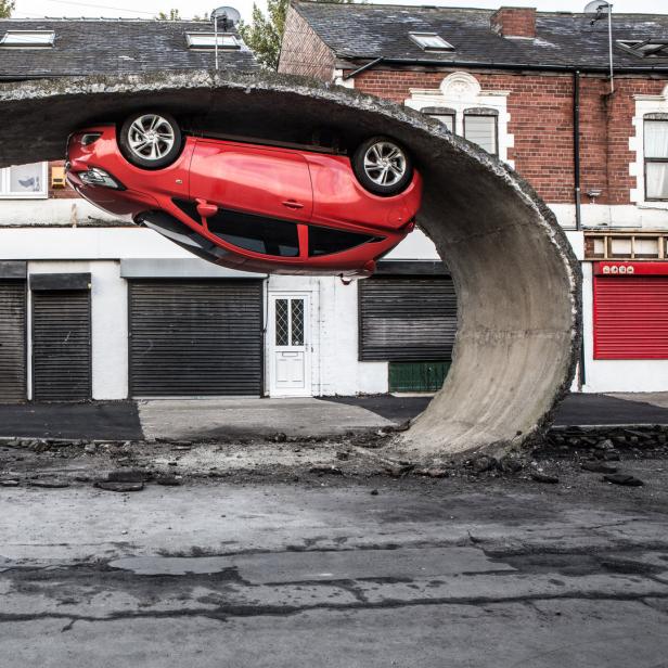 Ein rotes Auto steckt in einem gebogenen Betonstück vor einer Ladenzeile, ein Mann steht davor.