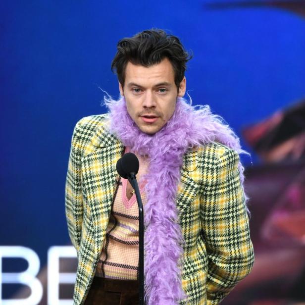 Harry Styles mit lila Boa und kariertem Sakko am Mikrofon.