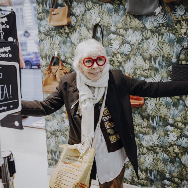 Eine ältere Frau mit roter Brille steht in einem Geschäft mit Taschen und einem „Styling Area!“-Schild.