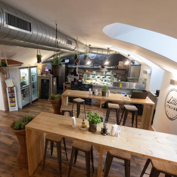 Innenansicht eines „Little Italian Deli“-Geschäfts mit Tischen und einer Theke.