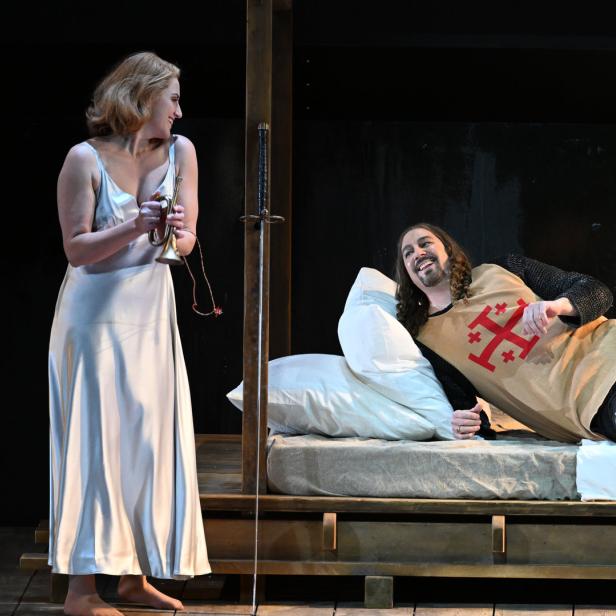 Eine Szene aus einem Theaterstück mit einer Frau mit Trompete und einem Ritter im Bett.