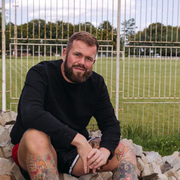 Ein bärtiger Mann mit Tätowierungen sitzt vor einem Fußballplatz auf einem Steinhaufen.