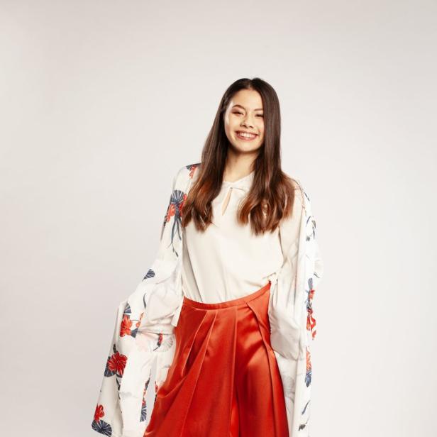 Eine lächelnde Frau posiert in einer weißen Bluse, einer floralen Jacke und einer roten Hose vor einem weißen Hintergrund.