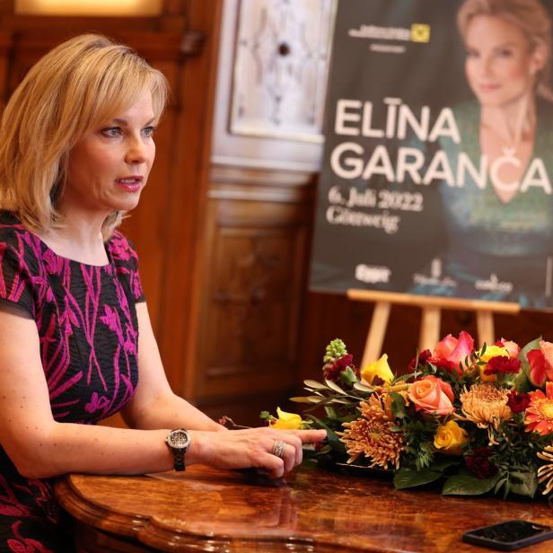 Eine Frau sitzt an einem Tisch mit Blumen; im Hintergrund ein Plakat von Elīna Garanča.