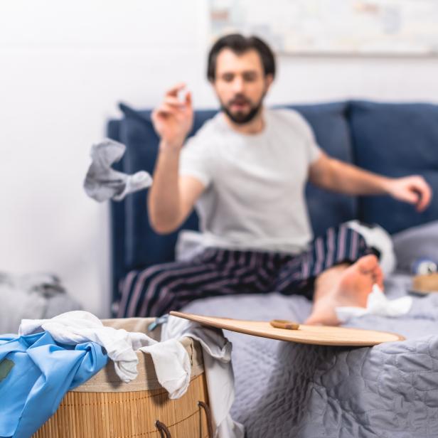 Ein Mann wirft Wäsche, während er auf einem unordentlichen Bett sitzt.