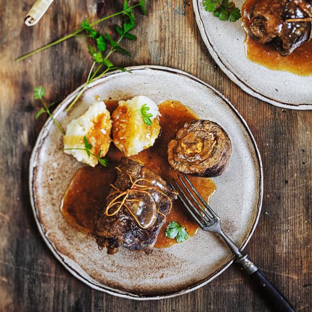 Zwei Teller mit Rinderrouladen, Kartoffelbeilagen und Soße auf einem Holztisch.