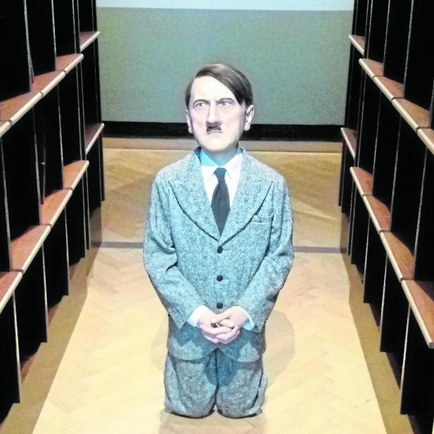 Eine Wachsfigur von Adolf Hitler kniet zwischen leeren Bücherregalen.