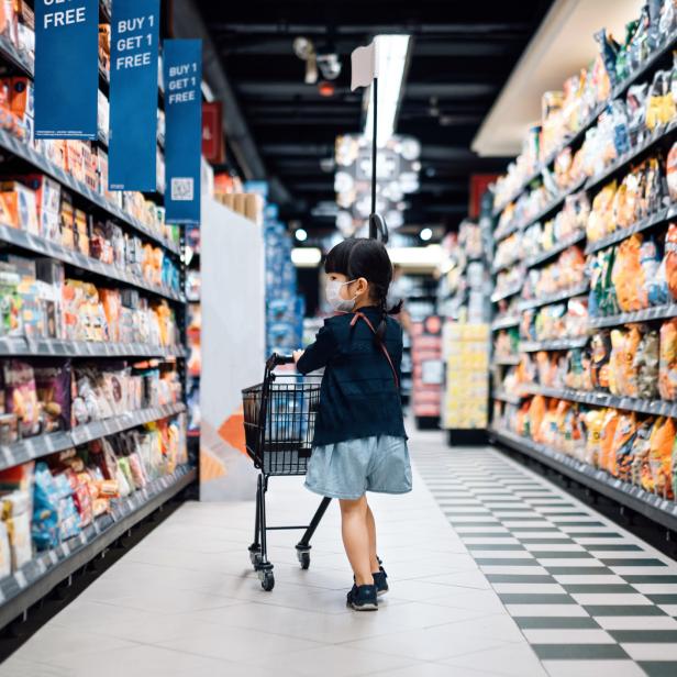 Ein kleines Mädchen mit Maske schiebt einen Einkaufswagen durch einen Supermarkt.