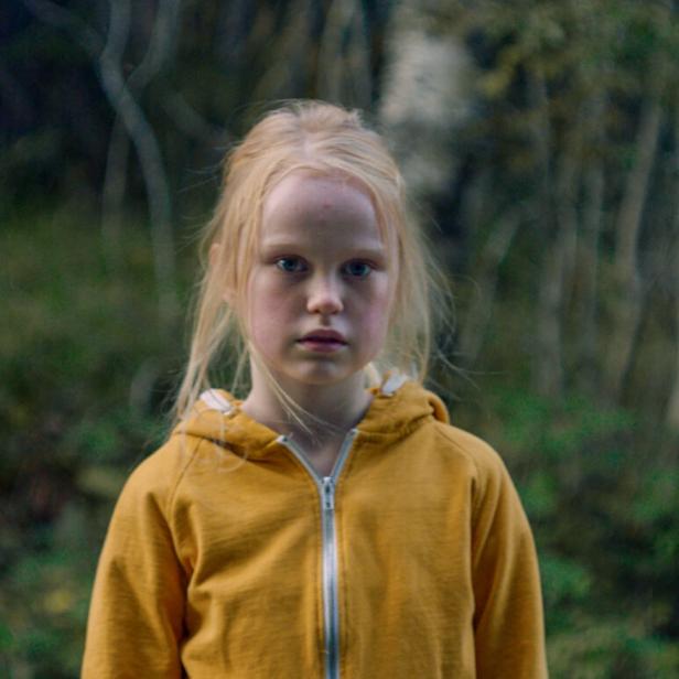 Ein junges Mädchen mit blonden Haaren und gelbem Kapuzenpullover steht vor einem Wald.