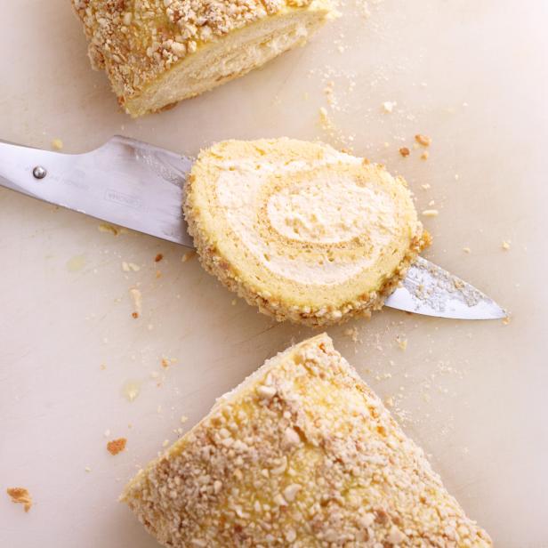 Ein angeschnittener Biskuitrollen-Kuchen mit Nussstreuseln liegt auf einem Schneidebrett.