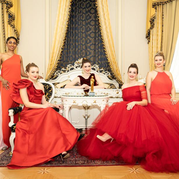 Fünf Frauen in roten Kleidern posieren in einem opulenten Raum mit goldenen Akzenten.