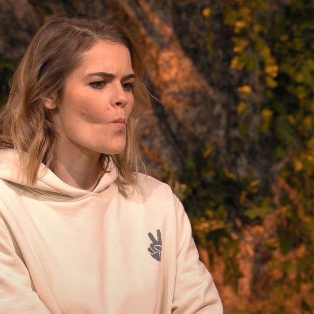 Eine Frau mit blonden Haaren und einem Kapuzenpullover mit Friedenszeichen vor einem Felsen.