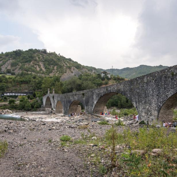 Eine alte Steinbrücke überspannt einen Fluss, an dem sich Menschen aufhalten.