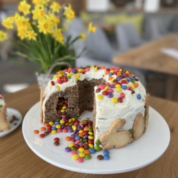 Ein angeschnittener Osterkuchen mit bunten Schokolinsen und Hasenkeksen steht auf einem Tisch.