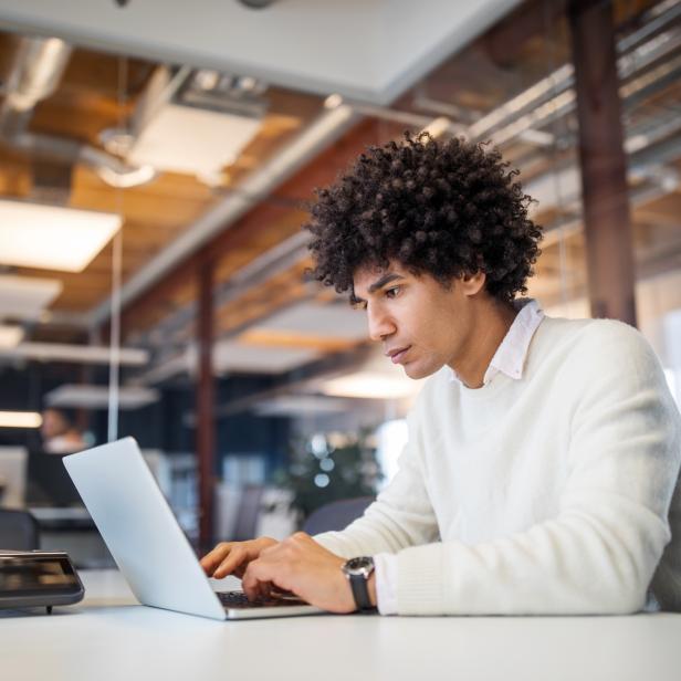 Ein junger Mann mit Afro sitzt an einem Schreibtisch und arbeitet am Laptop.