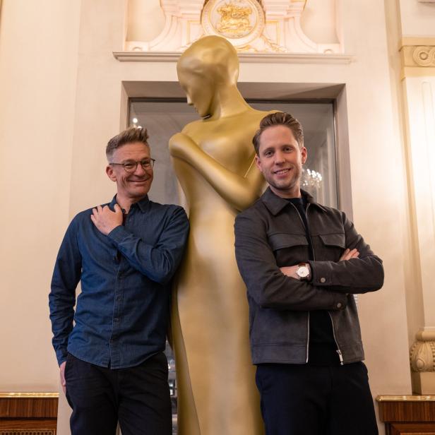Zwei Männer posieren neben einer goldenen Statue in einem eleganten Raum.