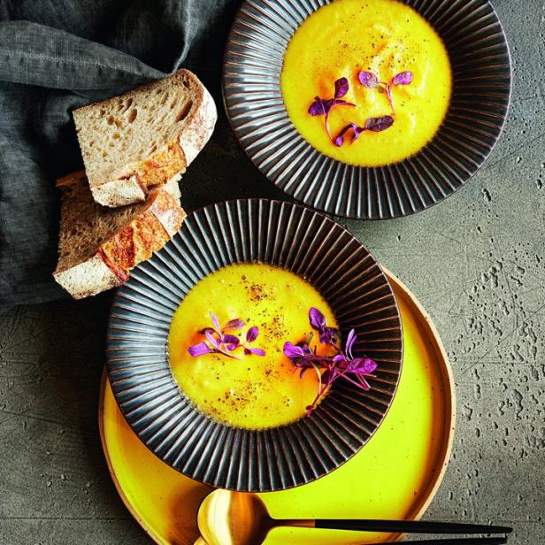 Zwei Teller mit gelber Suppe, garniert mit lila Kräutern, und Brot auf einem Tisch.