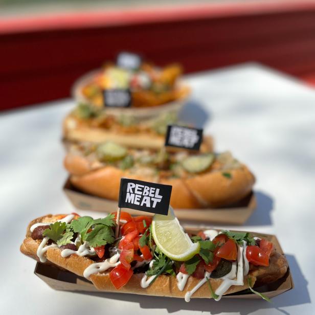Drei Hotdogs von „Rebel Meat“ mit verschiedenen Toppings werden auf einem Tisch präsentiert.