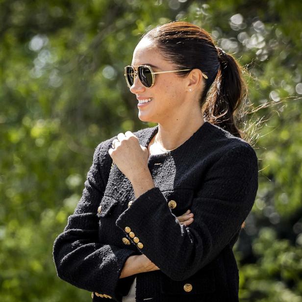 Meghan Markle lächelt mit verschränkten Armen und Sonnenbrille vor einem grünen Hintergrund.