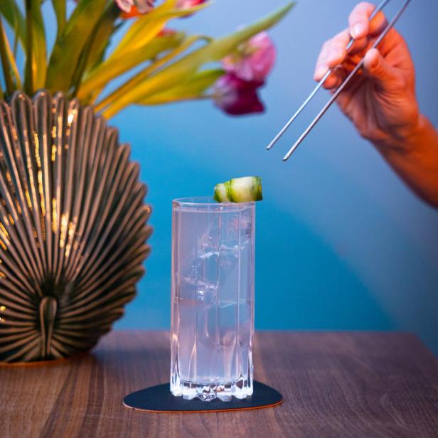 Ein Cocktail wird mit einer Gurkenscheibe garniert, im Hintergrund ein Blumenstrauß.