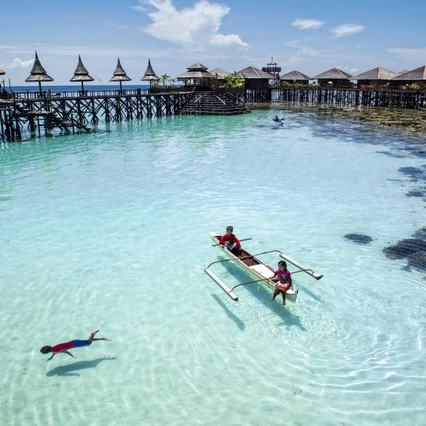 Kinder spielen im türkisfarbenen Wasser vor einer malerischen Insel mit Stelzenhäusern.