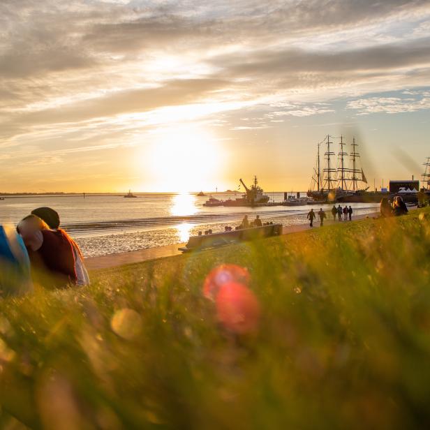 Menschen sitzen am Ufer und beobachten den Sonnenuntergang über dem Meer mit Segelschiffen.